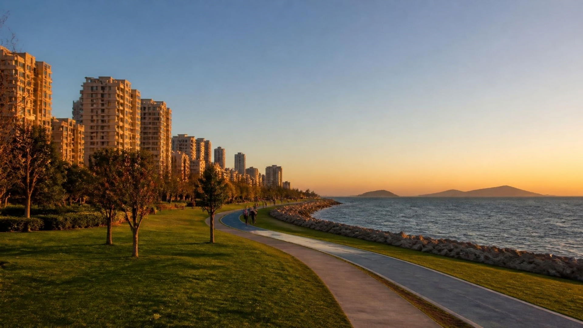 Seaside park path and apartment towers at sunset, with islands on the horizon